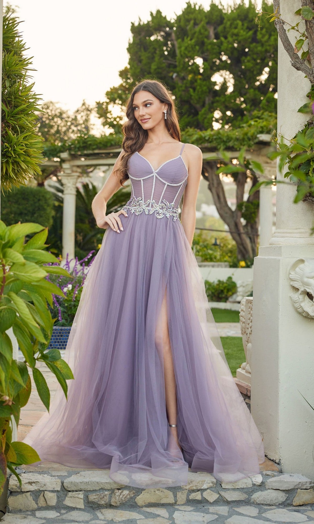 A woman stands outdoors in a lush garden wearing the Long Prom Dress: Juliet JT2638G in grey purple, featuring a corset bodice, sheer tulle skirt with silver embellishments, and a high slit.
